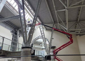 High-level industrial cleaning and maintenance work being carried out inside a modern building with steel beams and glass roof, using elevated work platforms and cherry pickers.