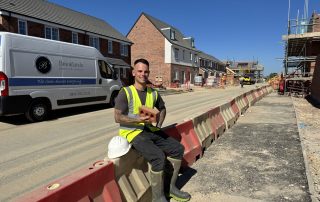 High-visibility worker sitting on barrier at a construction site with a brick in hand, blue sky background, showcasing outdoor construction and building maintenance services.