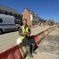 High-visibility worker sitting on barrier at a construction site with a brick in hand, blue sky background, showcasing outdoor construction and building maintenance services.