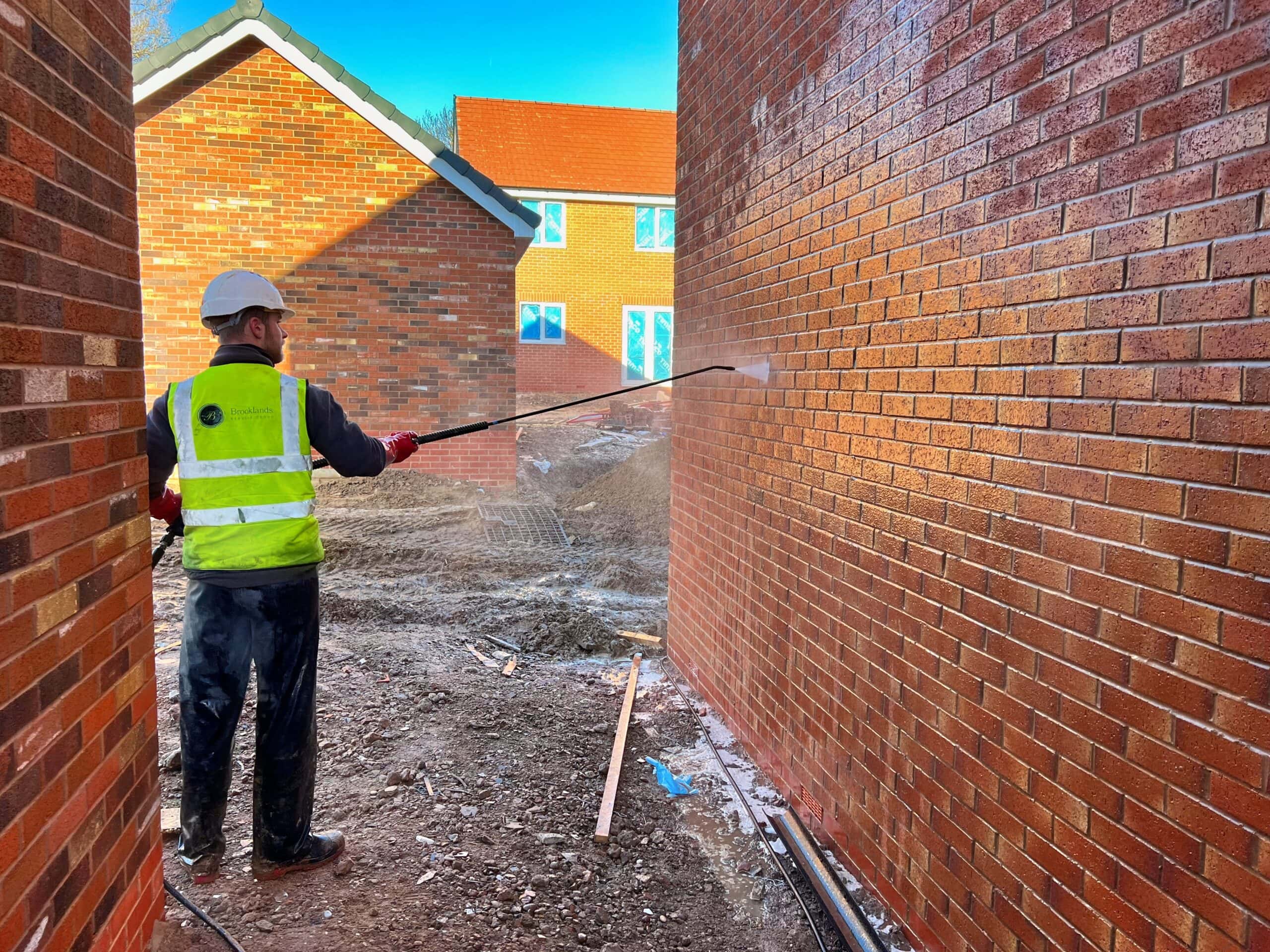 Man in high-visibility vest and helmet using pressure washer on brick walls.