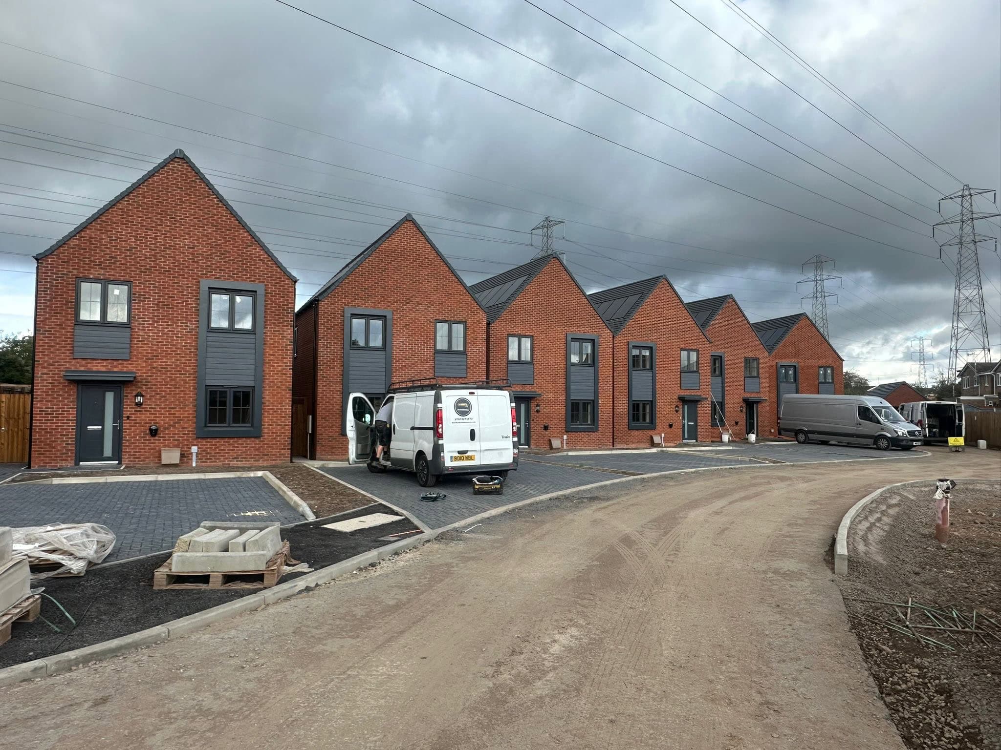 Newly built red brick houses with dark grey window frames and doors, under construction with landscaping work ongoing, parked vans with Brick Cleaning team on site, and a cloudy sky in the background.