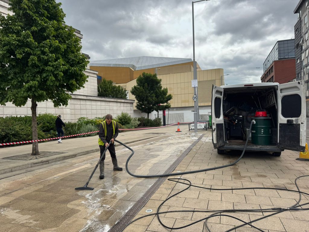 High-pressure surface cleaning by a professional with a vacuum system on a paved city walkway, with city buildings and cloudy sky in the background. Cleaning to remove oil spill from paved aras and streets.