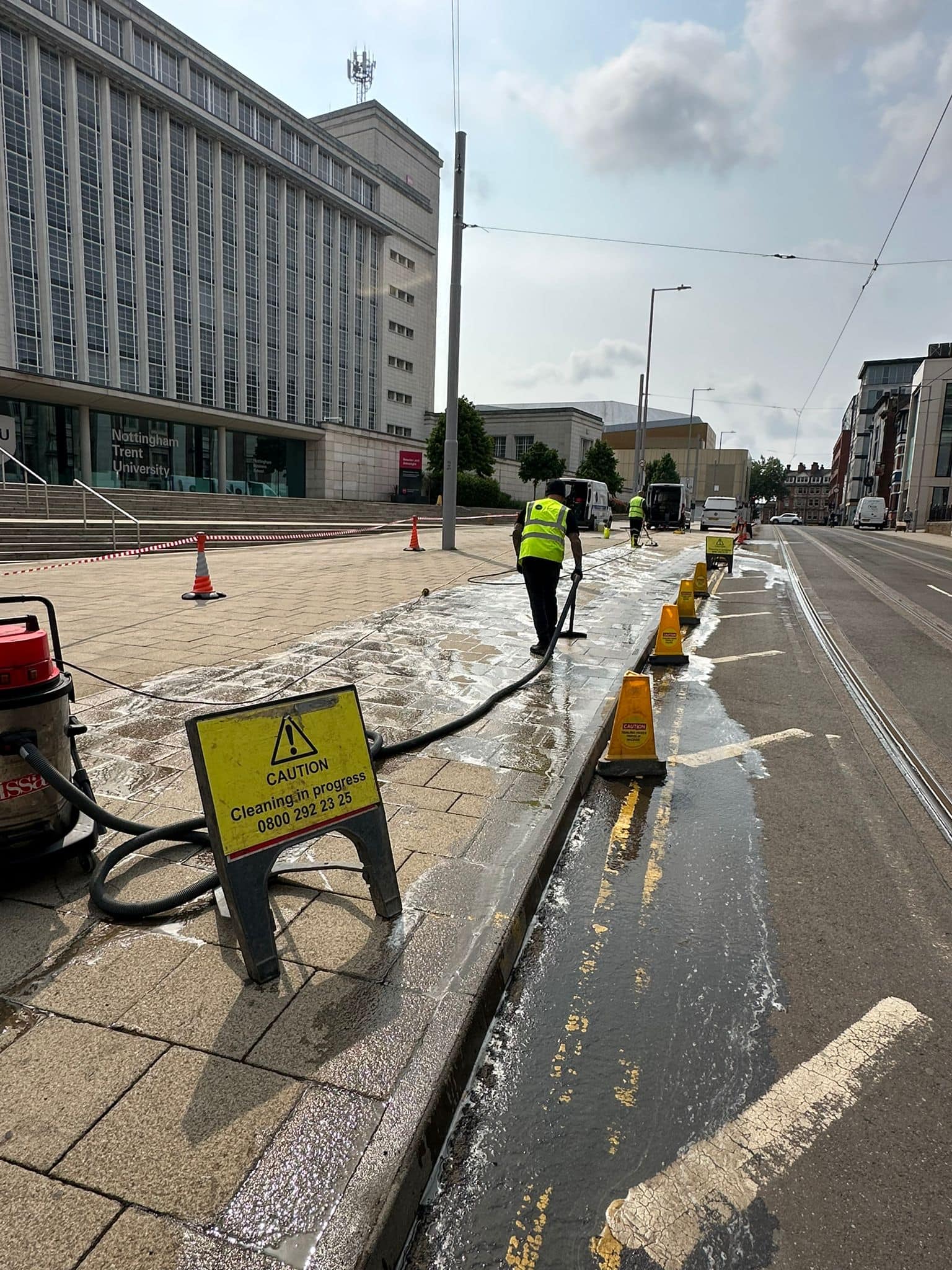 High-pressure cleaning of city pavement with workers in high visibility vests using hoses, outdoor cleaning equipment, and safety cones for tram tracks and pedestrian areas.