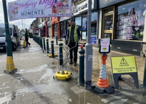 Deep cleaning of public pavement using high-powered pressure washer by professional team, with safety cones and signage, on a busy street with commercial shops and vehicles in the background.