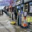 Deep cleaning of public pavement using high-powered pressure washer by professional team, with safety cones and signage, on a busy street with commercial shops and vehicles in the background.