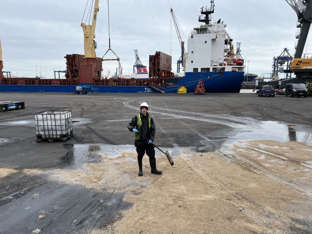 High-pressure cleaning on a harbour dock with a worker in safety gear, large ships, cranes, and industrial vehicles in the background.