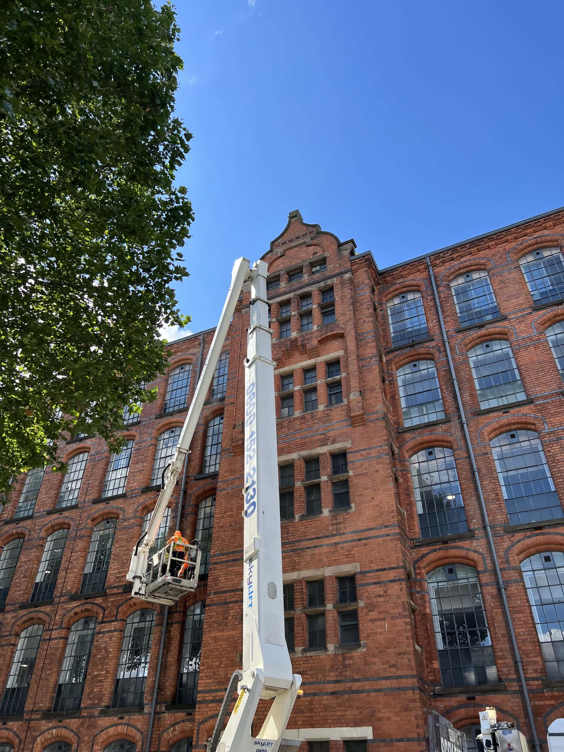 Elevated cherry picker cleaning historic red brick factory building with large windows.