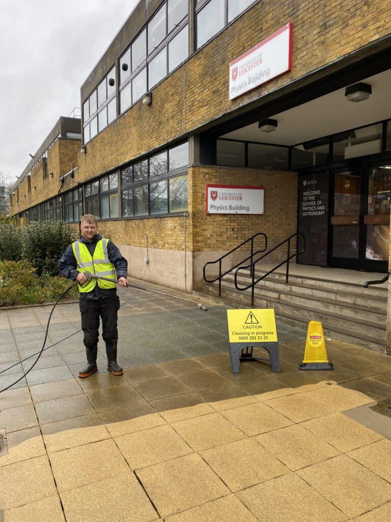 High-visibility worker pressure washing university building entrance, exterior cleaning service.