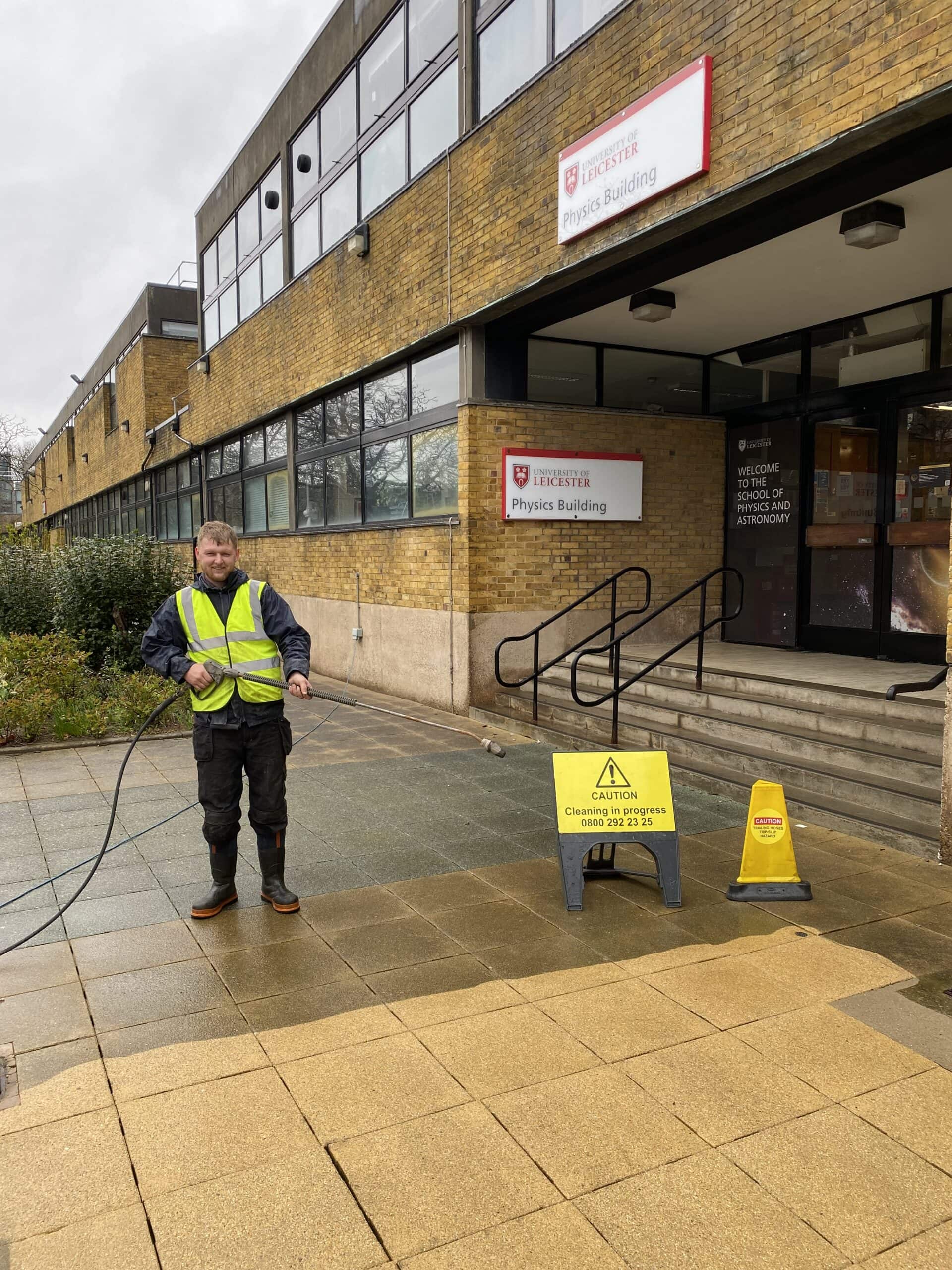 High-visibility worker pressure washing university building entrance, exterior cleaning service.