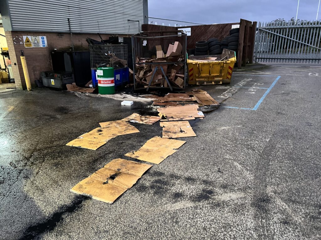 Spill on car park forecourt before cleaning. Overflowing waste bins with scattered cardboard and debris outside industrial facility.