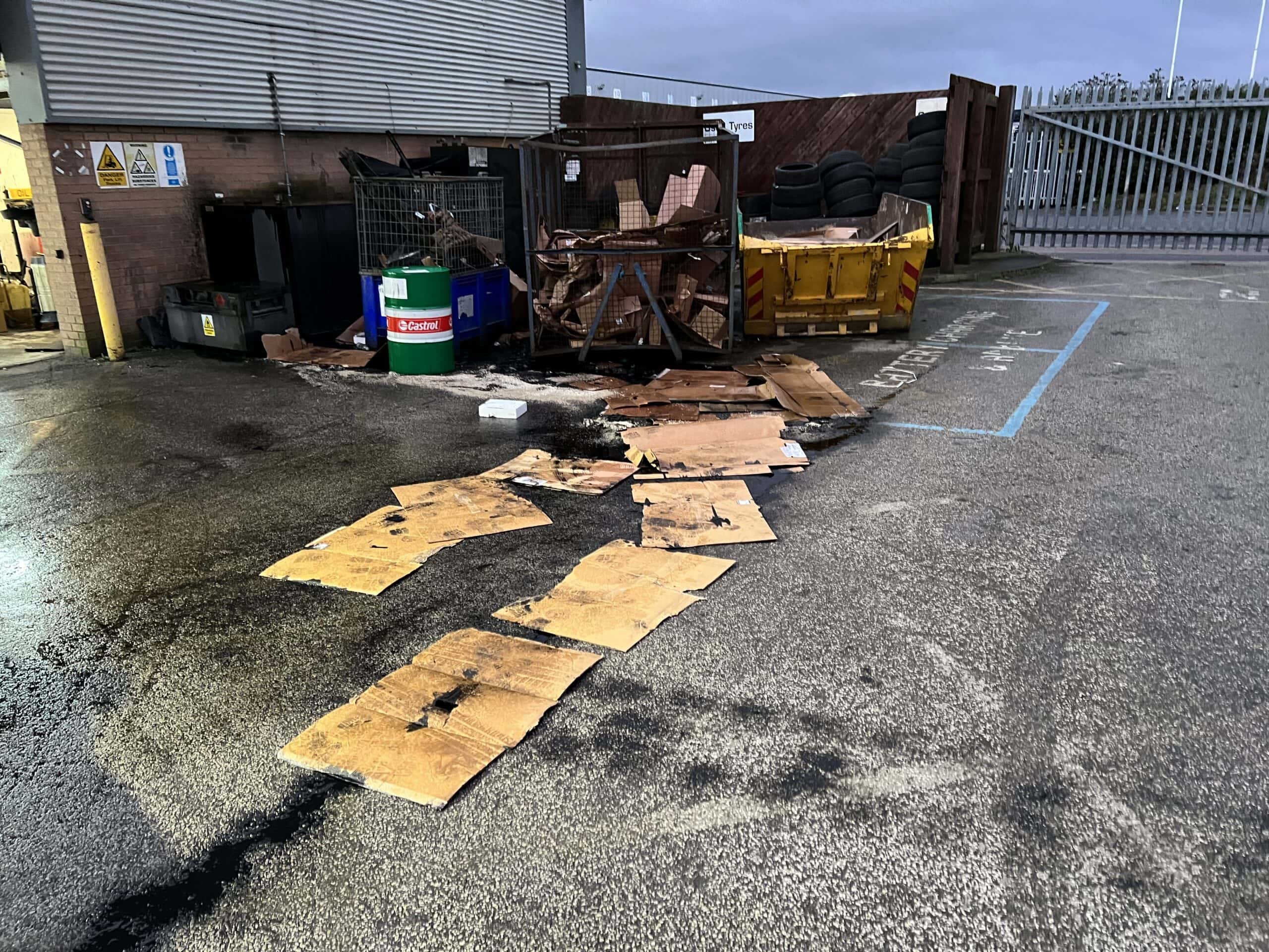 Spill on car park forecourt before cleaning. Overflowing waste bins with scattered cardboard and debris outside industrial facility.